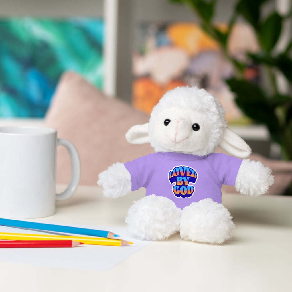 Plush white lamb wearing a purple shirt with "Loved by God" text sitting on a white table with colored pencils and a mug nearby