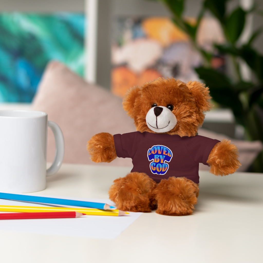 Brown teddy bear wearing a maroon shirt with "Loved by God" text sitting on a white table with colored pencils and a mug nearby