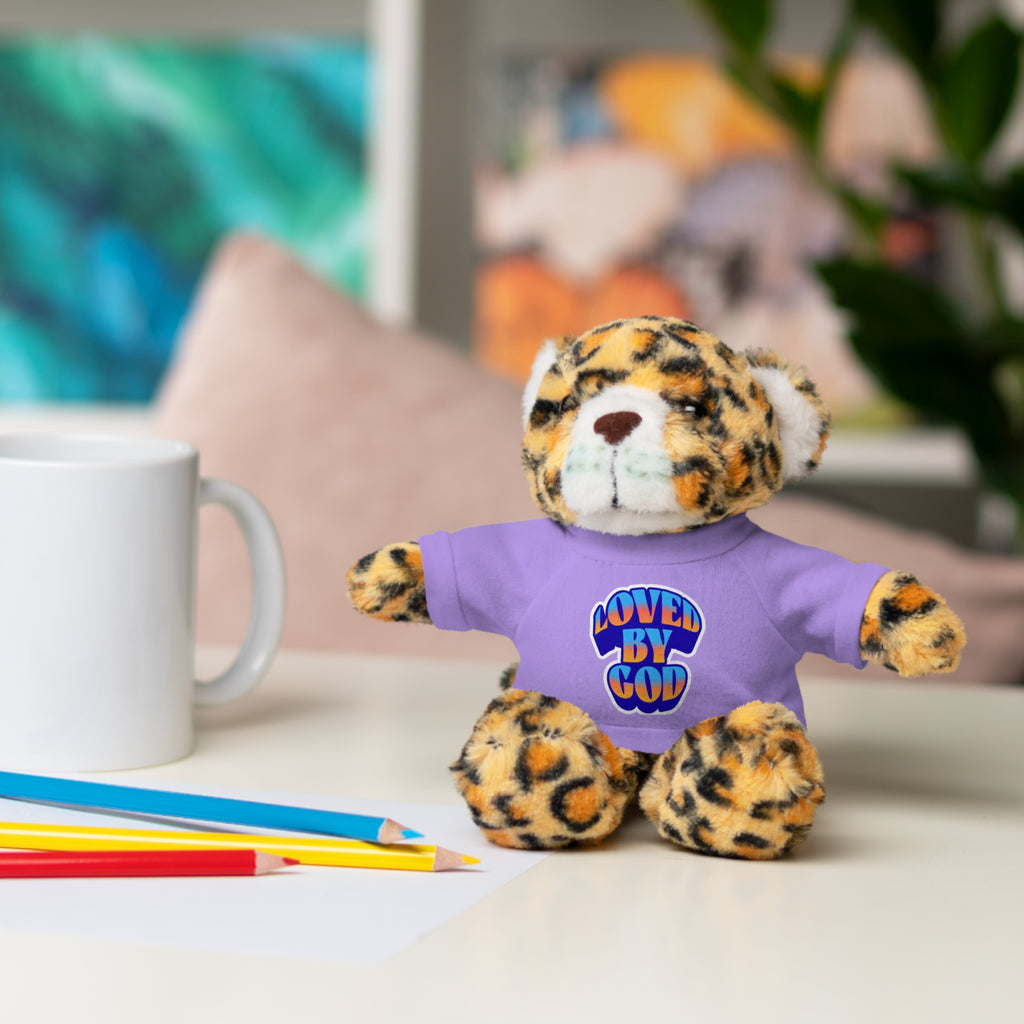 Stuffed leopard toy wearing purple shirt with "Loved by God" text sitting on desk near colored pencils and mug
