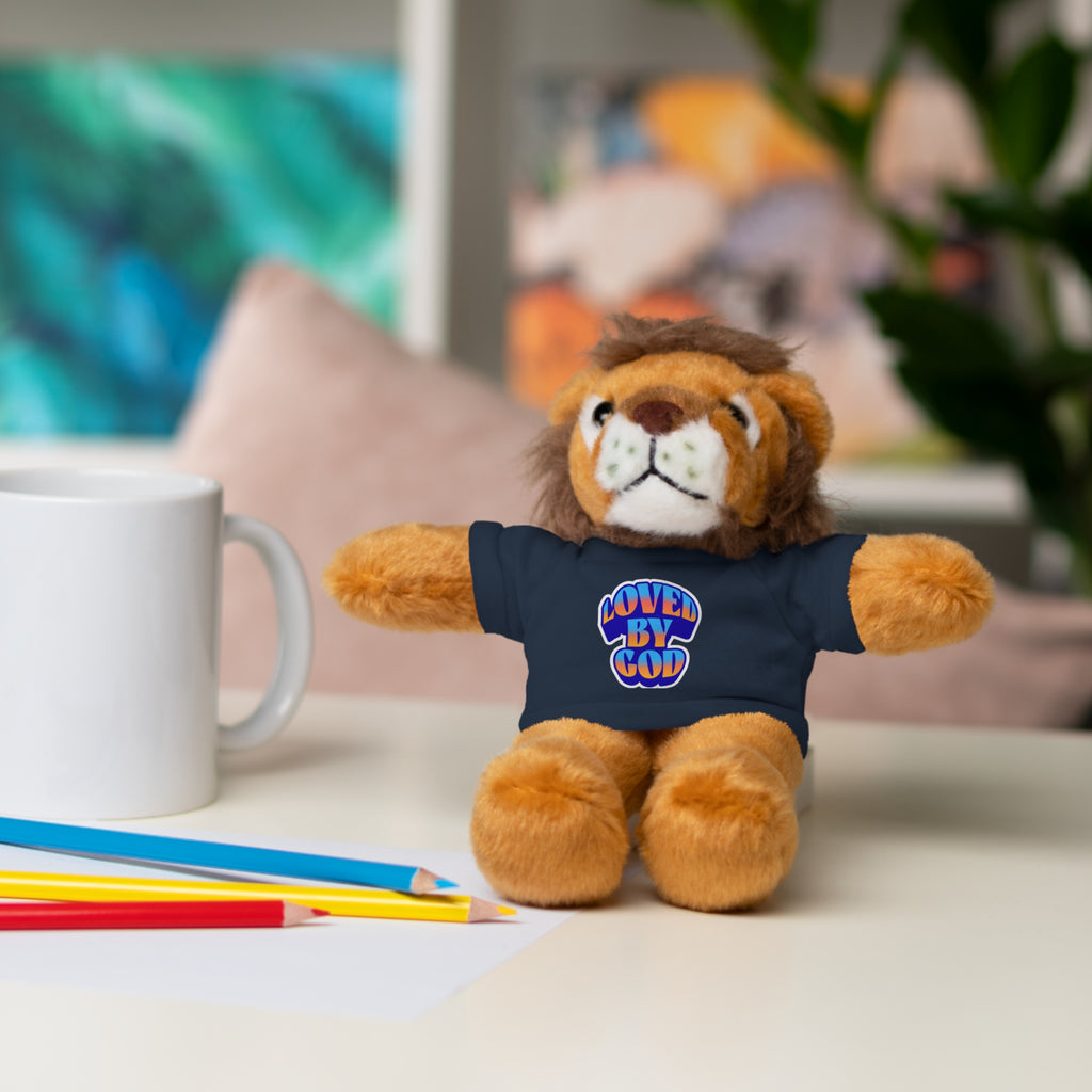 Plush lion toy wearing a navy blue shirt with loved by God text, sitting on a desk near colored pencils and a mug.
