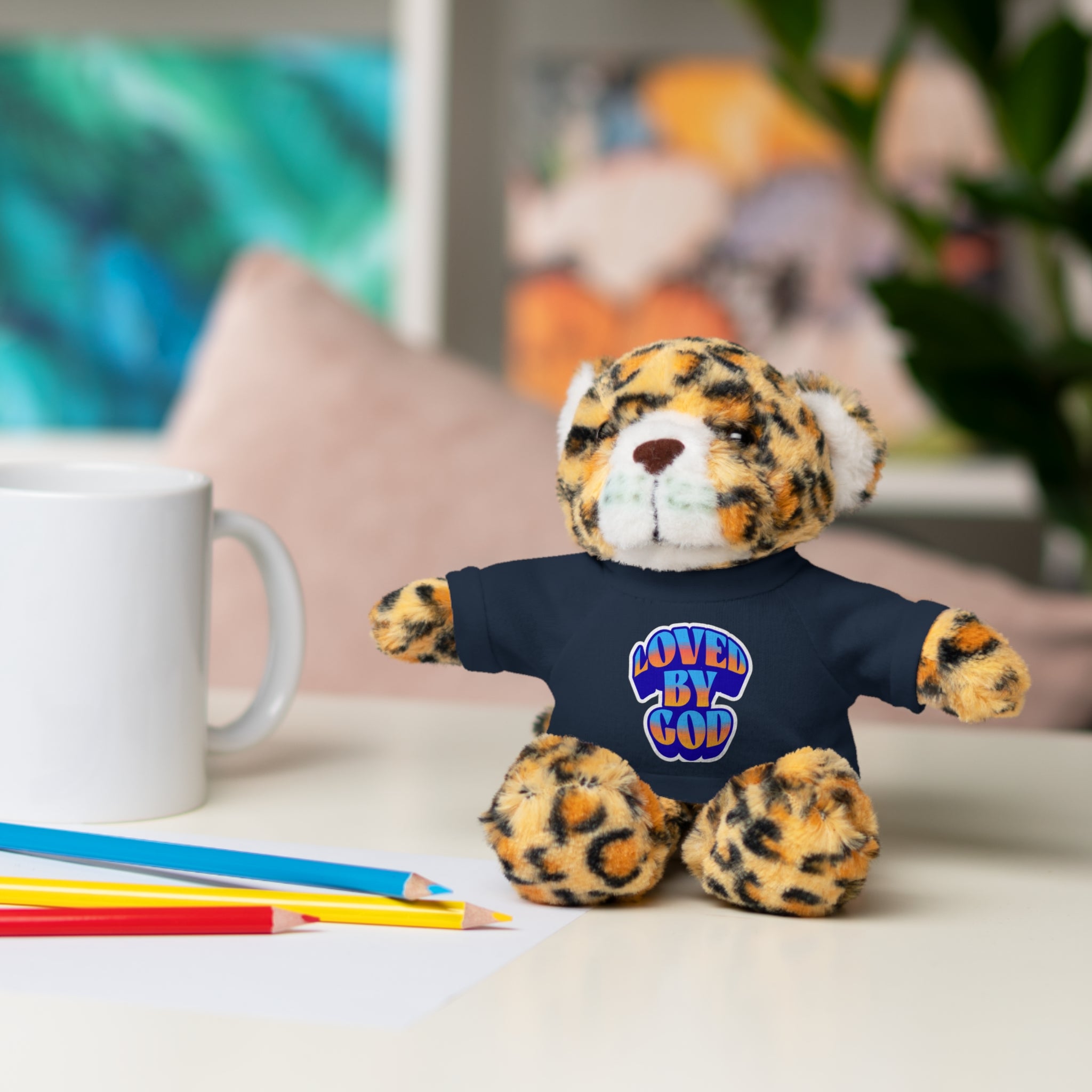 Small plush leopard toy wearing a black shirt with colorful "Loved by God" text sitting on a table near pencils and a white mug