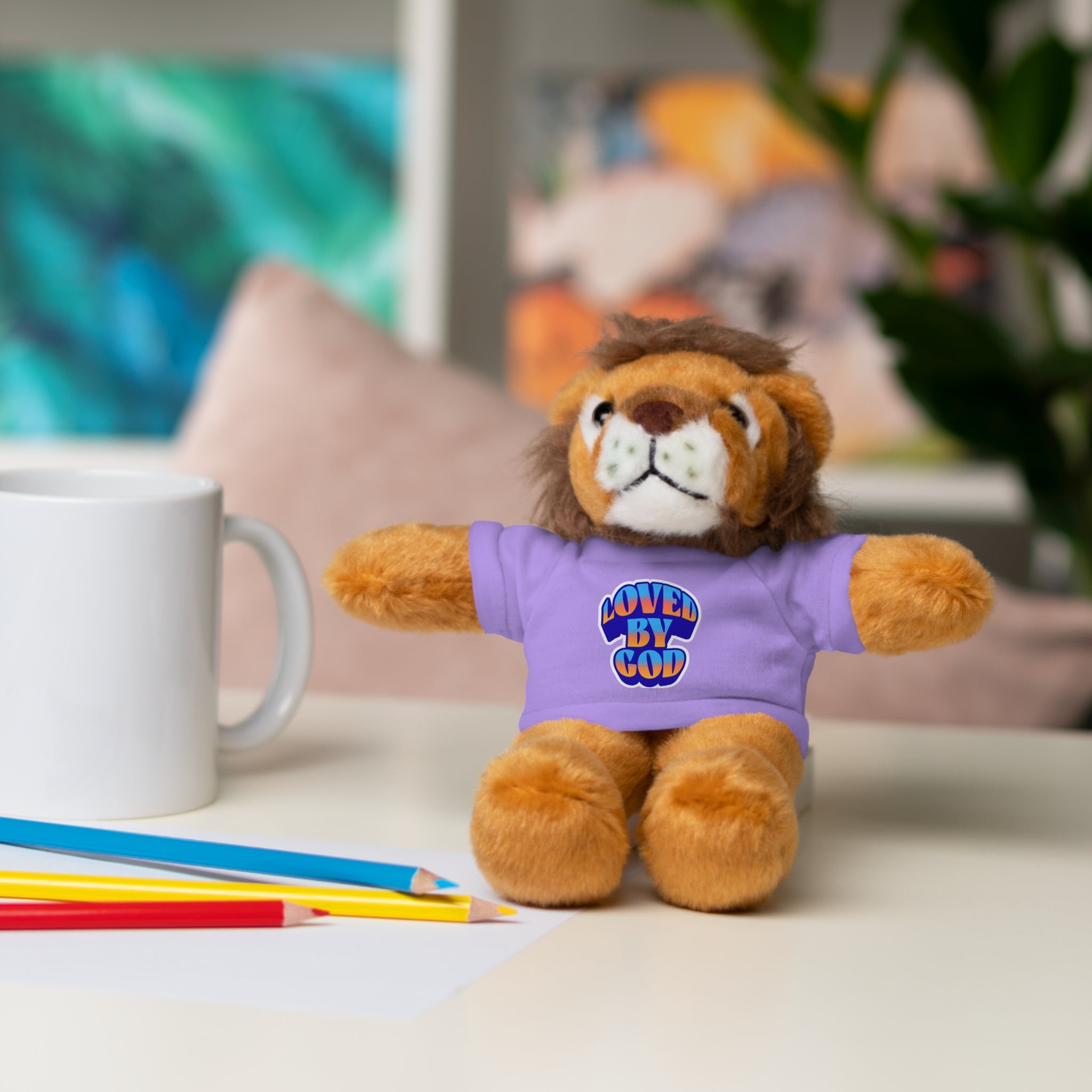 Plush lion toy wearing a purple "Loved by God" t-shirt sitting on a desk with pencils and a white mug nearby
