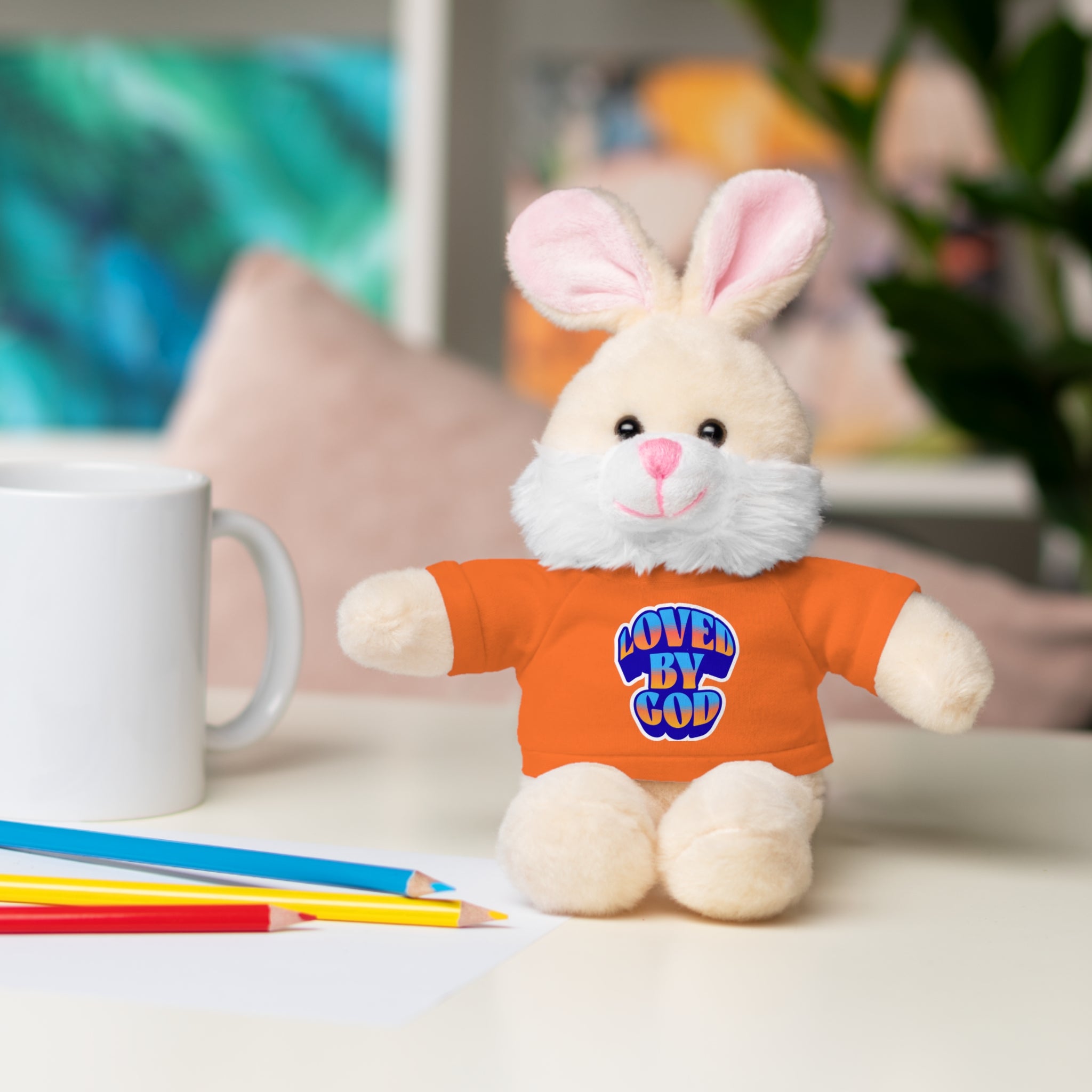 Soft plush bunny wearing an orange shirt with "Loved by God" text sitting next to colored pencils and a white mug.