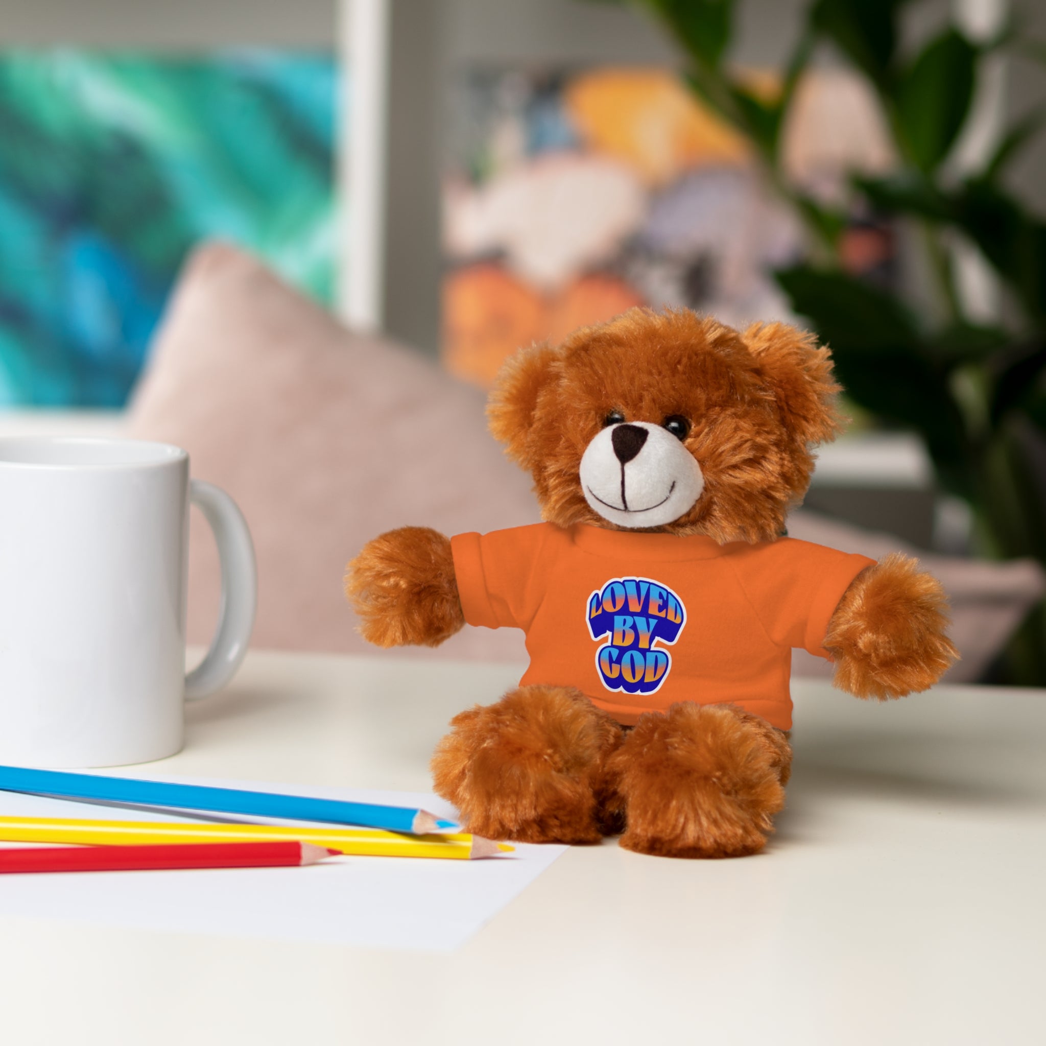 Brown teddy bear wearing an orange shirt with "Loved by God" text sitting on a white desk beside colored pencils and white mug