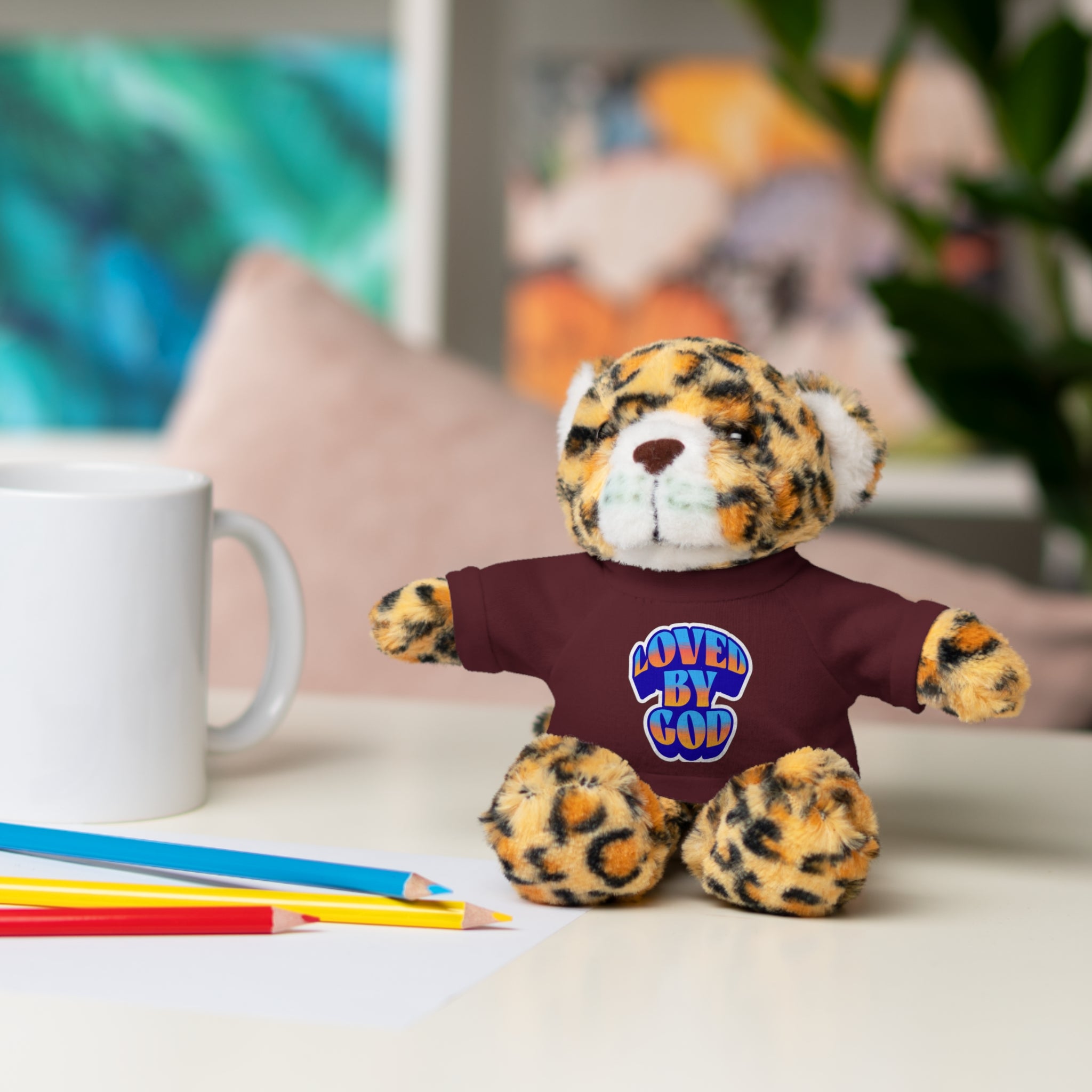 Plush leopard print teddy bear wearing a maroon shirt with "Loved by God" text sitting on a white table near colored pencils and a mug