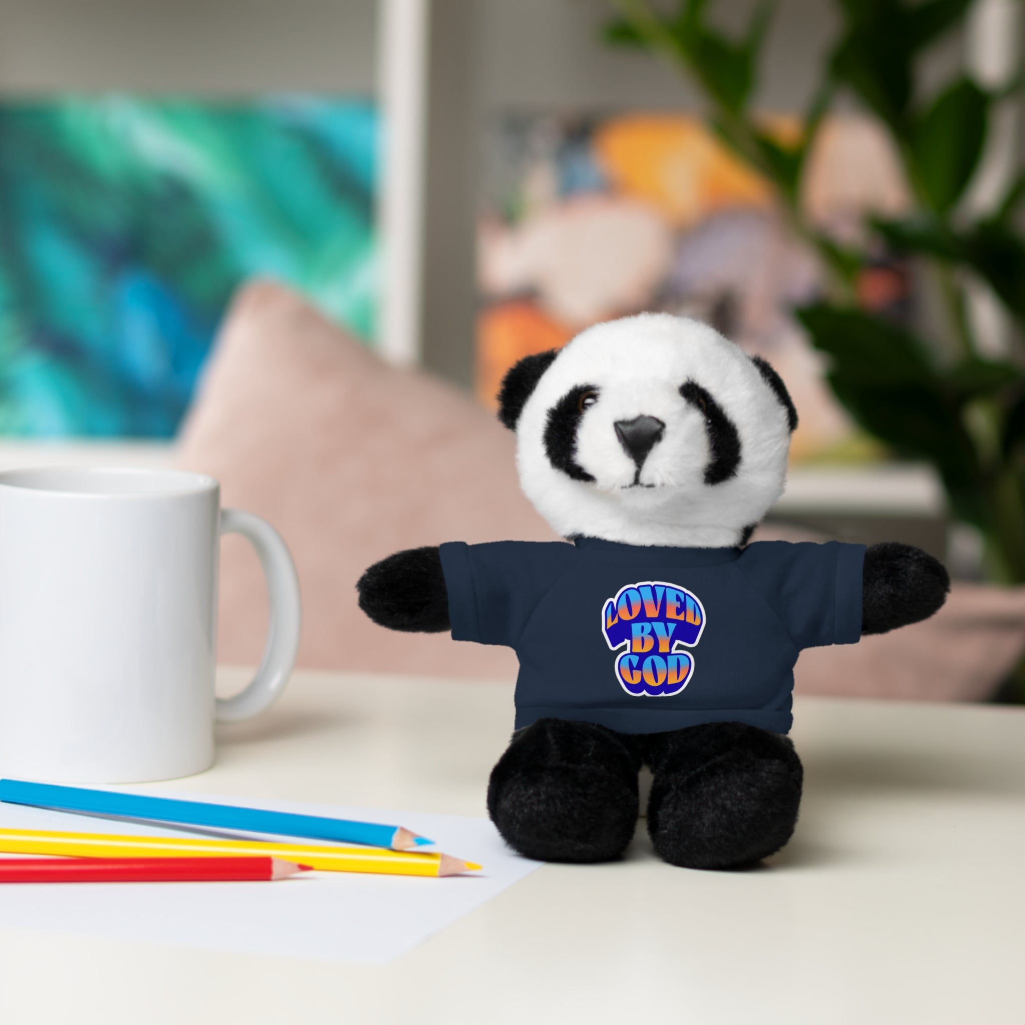 Small panda plush toy wearing a navy blue shirt with loved by God text sitting on a desk next to pencils and a mug