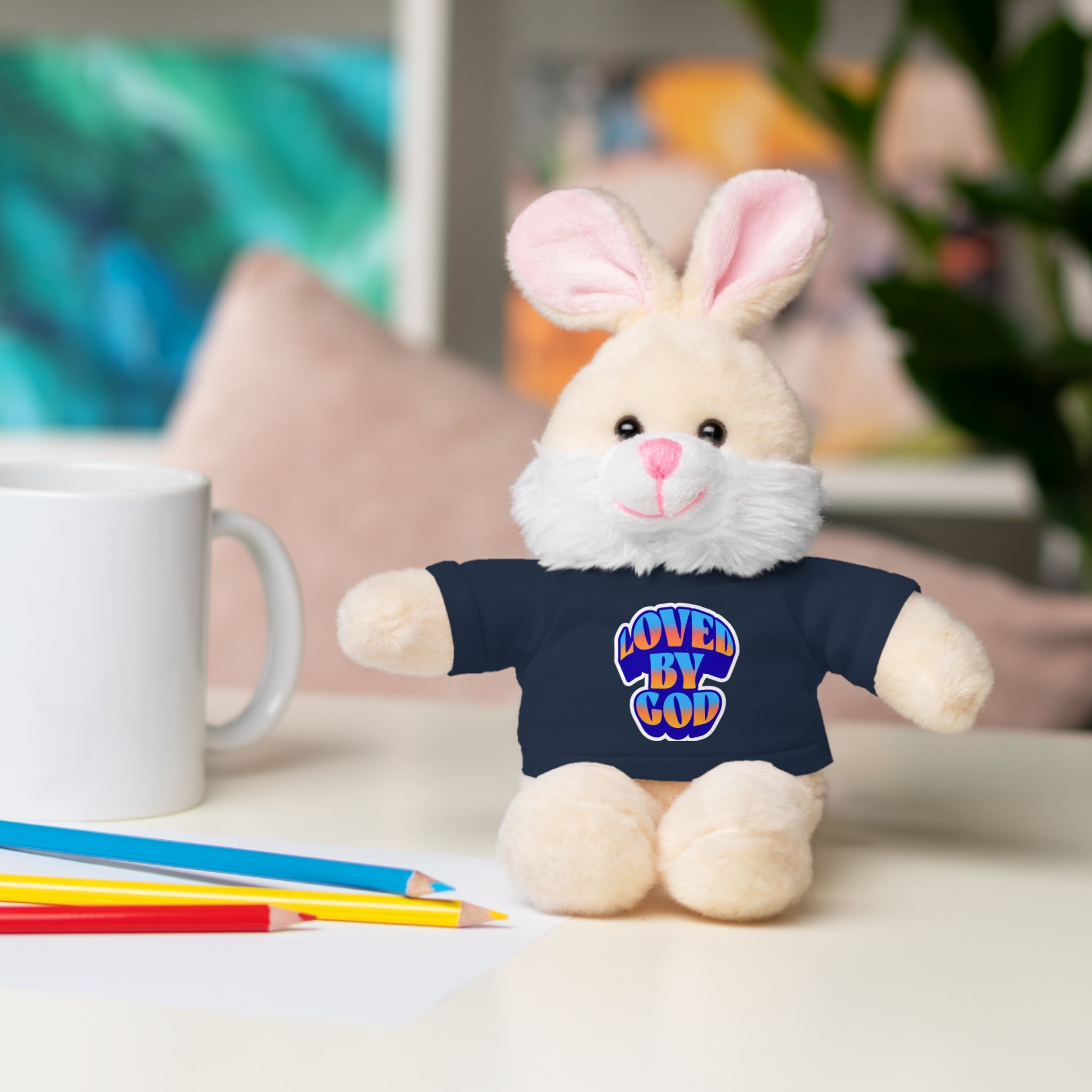 Plush bunny wearing a navy shirt with colorful "Loved by God" text sitting next to pencils and a white mug on a table.