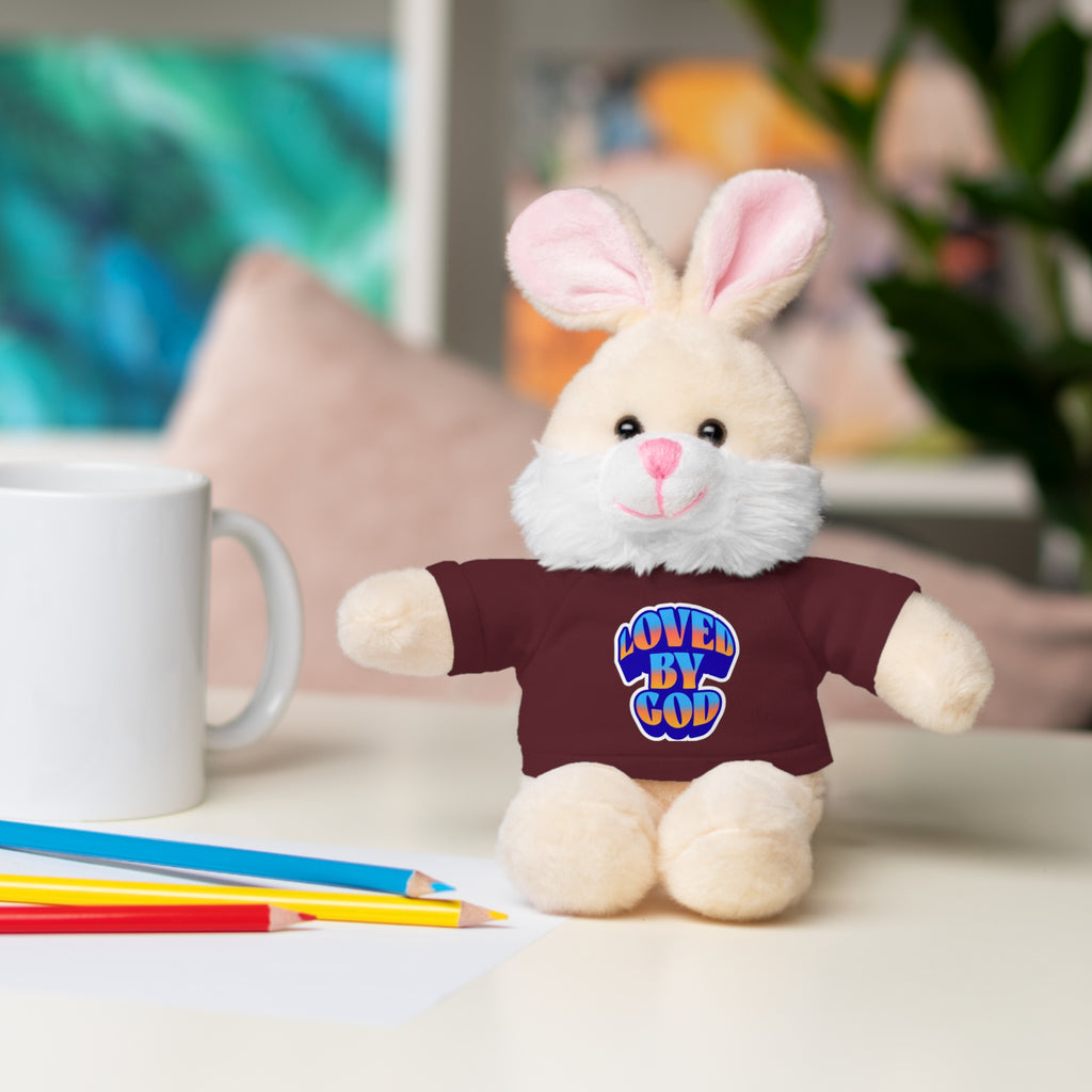 Cream-colored plush bunny wearing a maroon shirt with "Loved by God" text sitting on a desk next to colored pencils and a white mug
