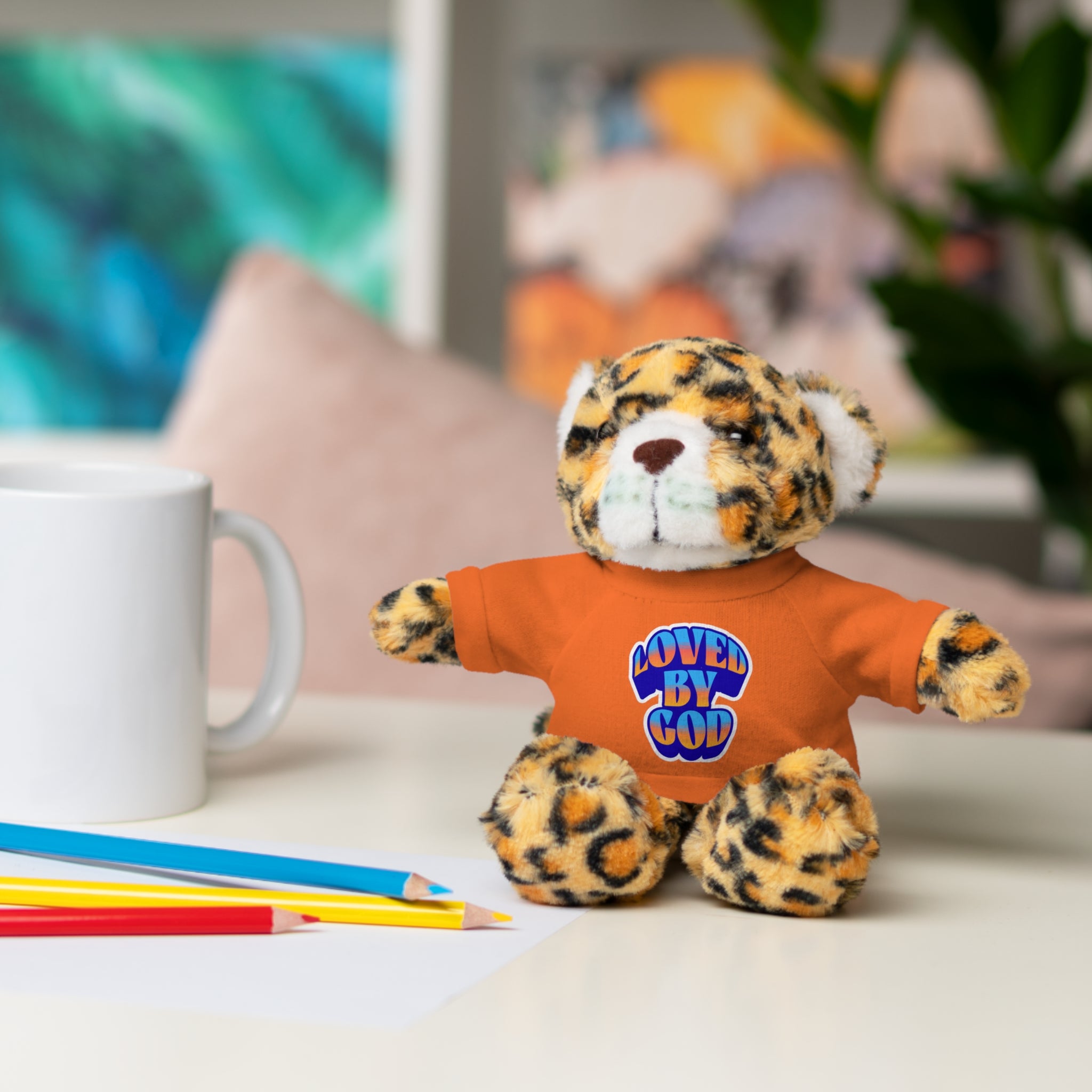 Plush leopard toy wearing orange "Loved By God" shirt sitting on desk with colored pencils and white mug nearby