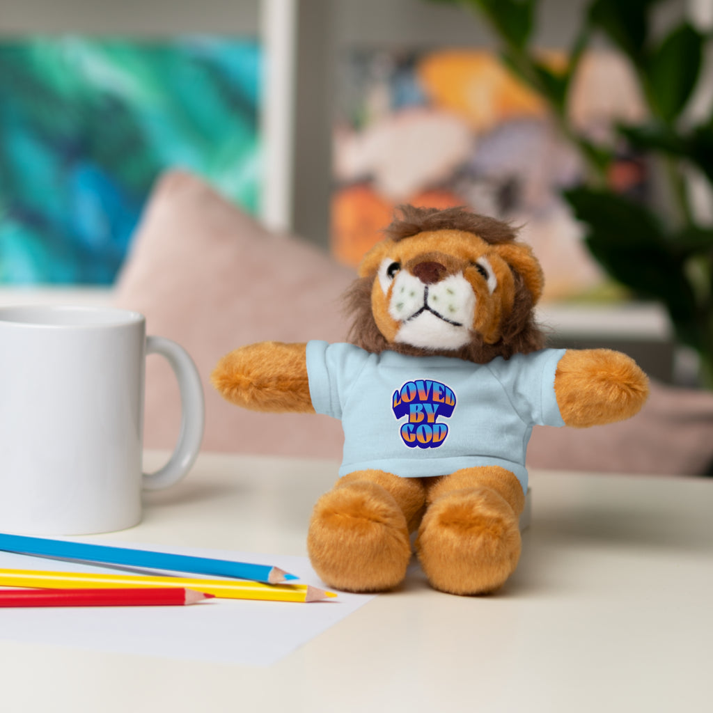 Plush lion toy wearing a light blue shirt with Loved by God text sitting on a desk next to colored pencils and a white mug.