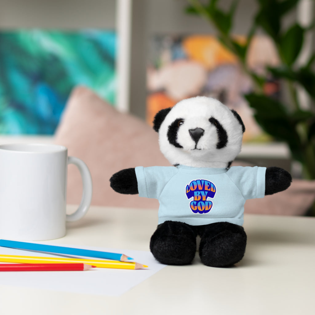 Small plush panda wearing a light blue shirt with "Loved by God" text sitting on a desk next to colored pencils and a white mug