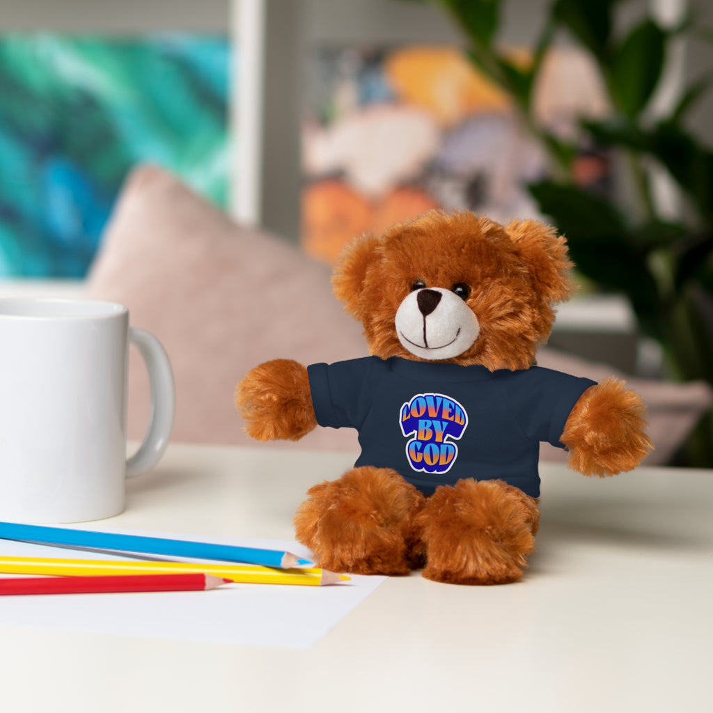 Brown teddy bear wearing navy blue shirt with Love By God logo sitting on white table next to colored pencils and white mug
