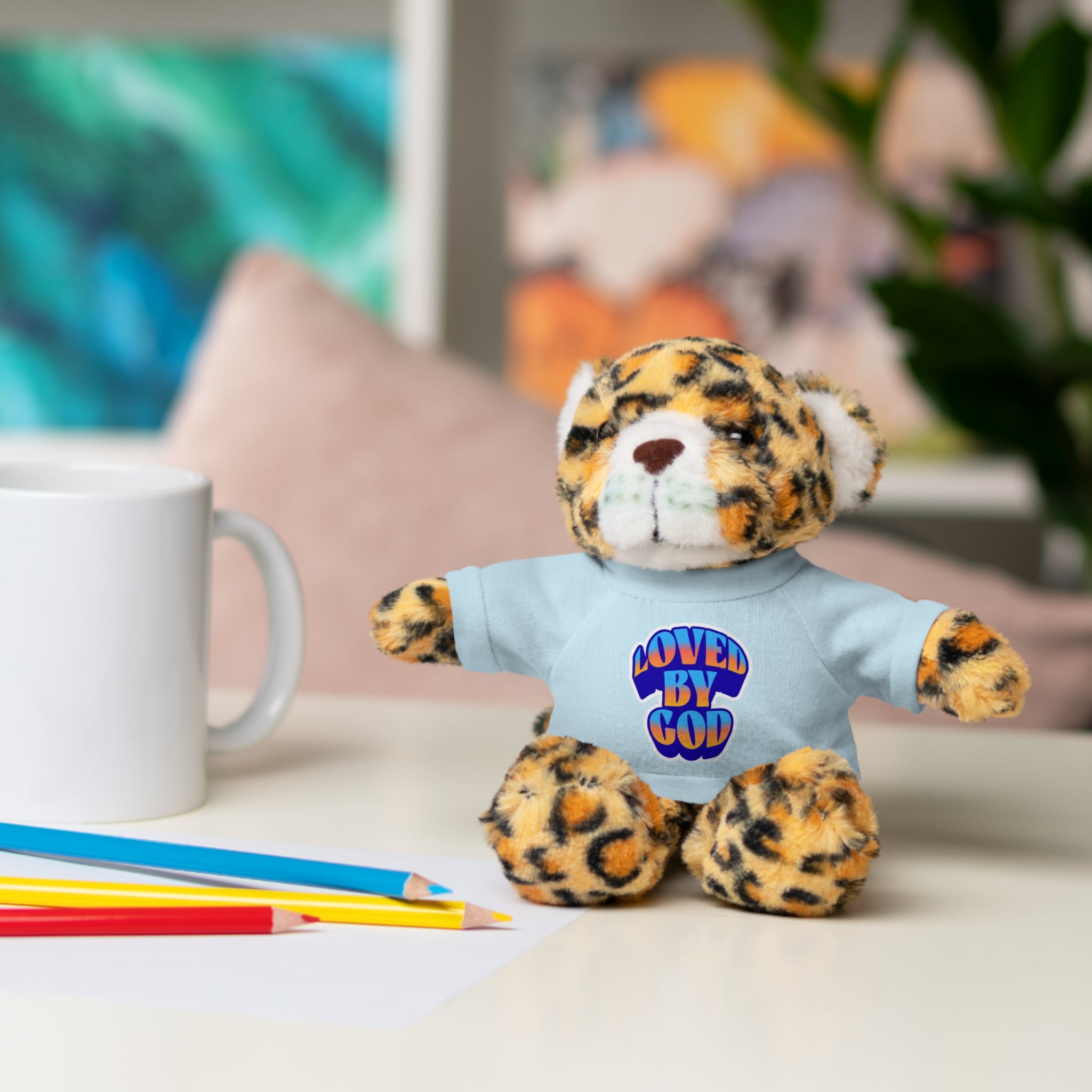 Small plush leopard toy wearing a blue shirt with "Loved by God" text sitting on a table next to pencils and a mug