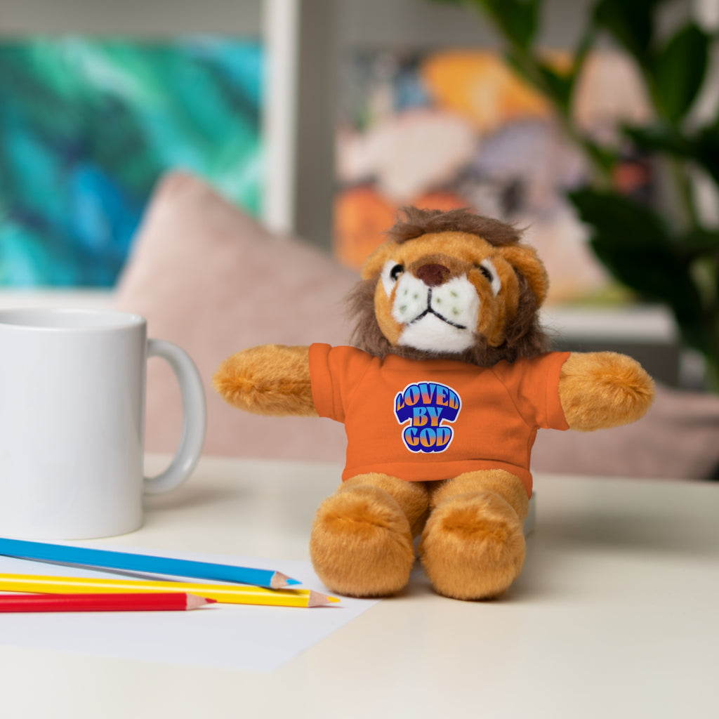 Plush lion toy wearing an orange shirt with Loved By God text sitting on a desk near pencils and a white mug