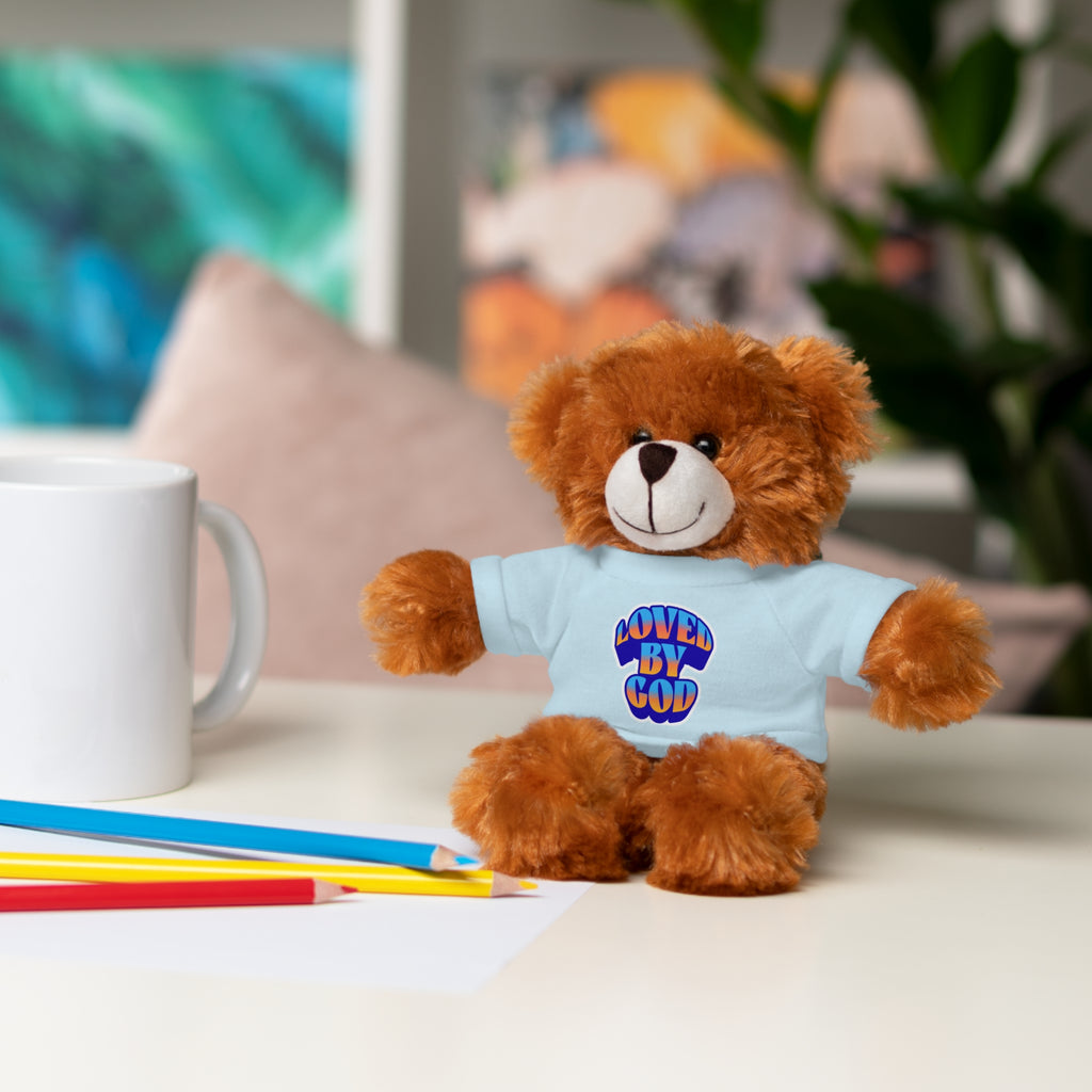Brown teddy bear wearing a white "Loved by God" shirt sitting on a table with colored pencils and a white mug nearby
