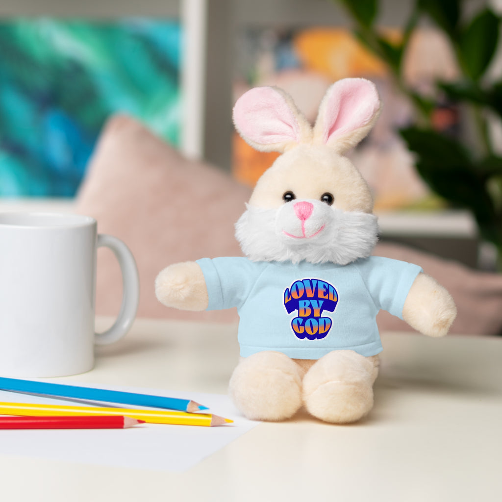 Plush bunny wearing a blue shirt with "Loved by God" text sitting on a table next to colored pencils and a white mug.